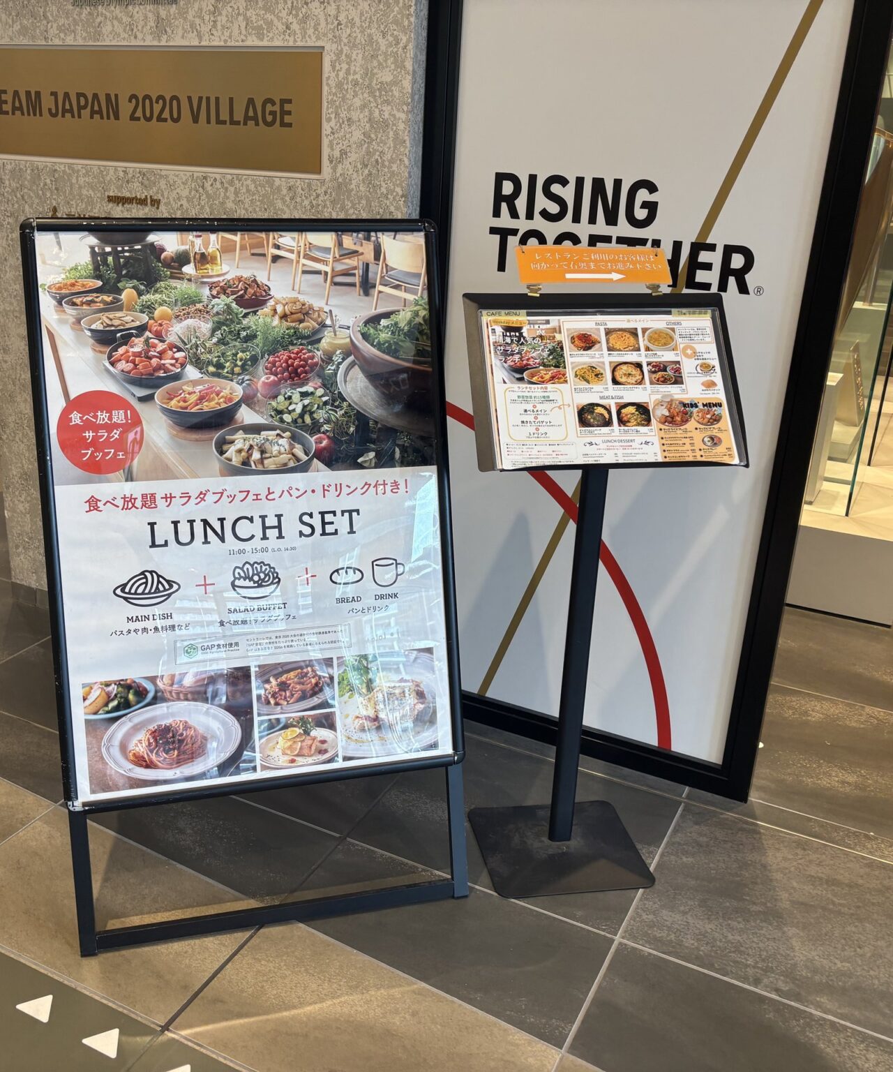 施設入口前に2点の案内物。左はA型スタンド看板で「食べ放題サラダブッフェとパン・ドリンク付き LUNCH SET 11:00〜15:00」と記載され、料理写真が掲載されている。右はスタンド式メニュー表。背後に「TEAM JAPAN 2020 VILLAGE」のプレートと「RISING TOGETHER」のサインが見える。
