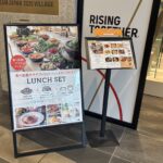 施設入口前に2点の案内物。左はA型スタンド看板で「食べ放題サラダブッフェとパン・ドリンク付き LUNCH SET 11:00〜15:00」と記載され、料理写真が掲載されている。右はスタンド式メニュー表。背後に「TEAM JAPAN 2020 VILLAGE」のプレートと「RISING TOGETHER」のサインが見える。
