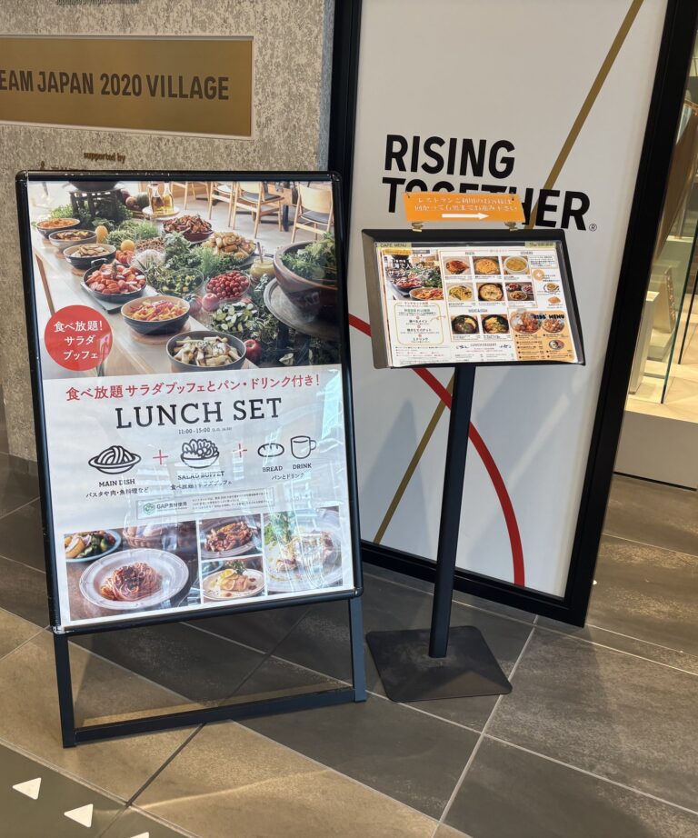 施設入口前に2点の案内物。左はA型スタンド看板で「食べ放題サラダブッフェとパン・ドリンク付き LUNCH SET 11:00〜15:00」と記載され、料理写真が掲載されている。右はスタンド式メニュー表。背後に「TEAM JAPAN 2020 VILLAGE」のプレートと「RISING TOGETHER」のサインが見える。