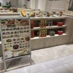 飲食店内、皿に載った料理の食品サンプルが並ぶ棚、下段に飲料のペットボトルと缶が陳列され、左手前にメニュー看板が置かれている。奥には座席と人物の一部が写っている。