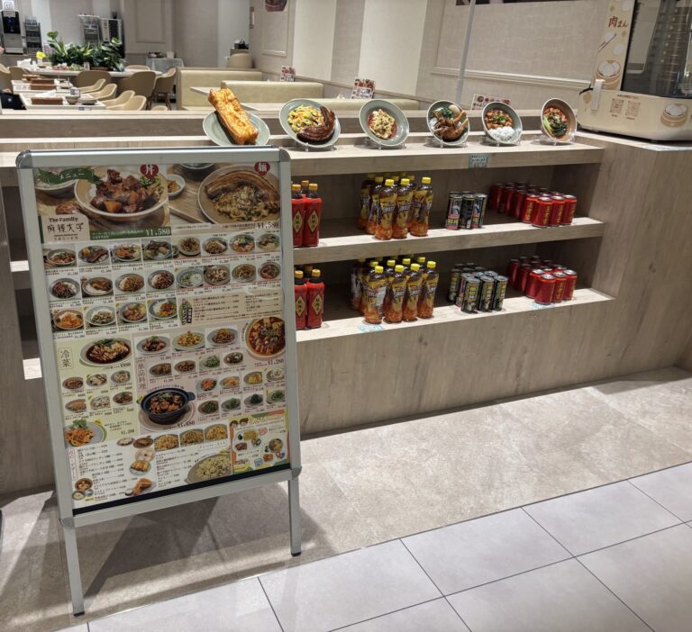 飲食店内、皿に載った料理の食品サンプルが並ぶ棚、下段に飲料のペットボトルと缶が陳列され、左手前にメニュー看板が置かれている。奥には座席と人物の一部が写っている。